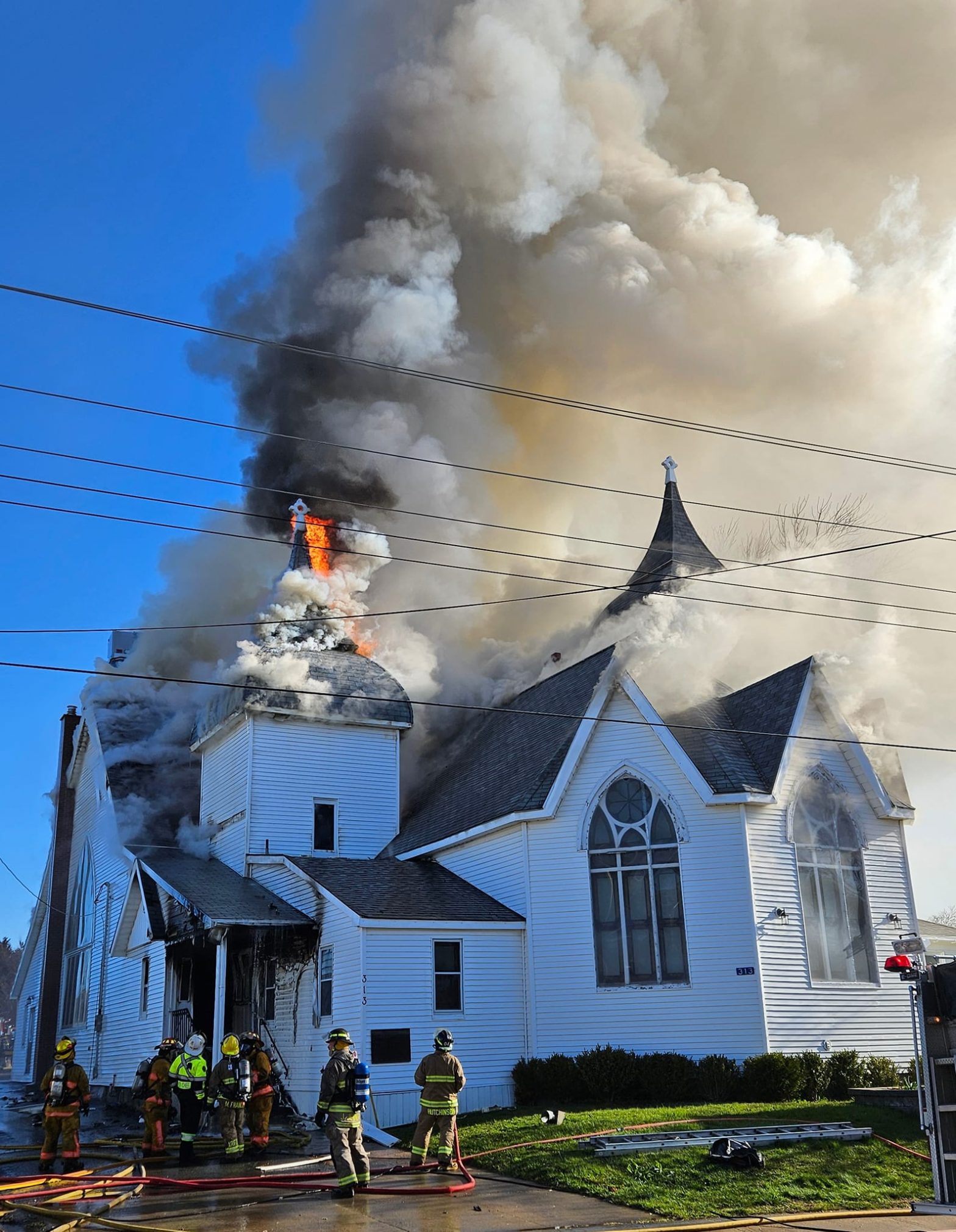 Flames and smoke emerge from large white wooden church as firefighters aim to control the blaze.
