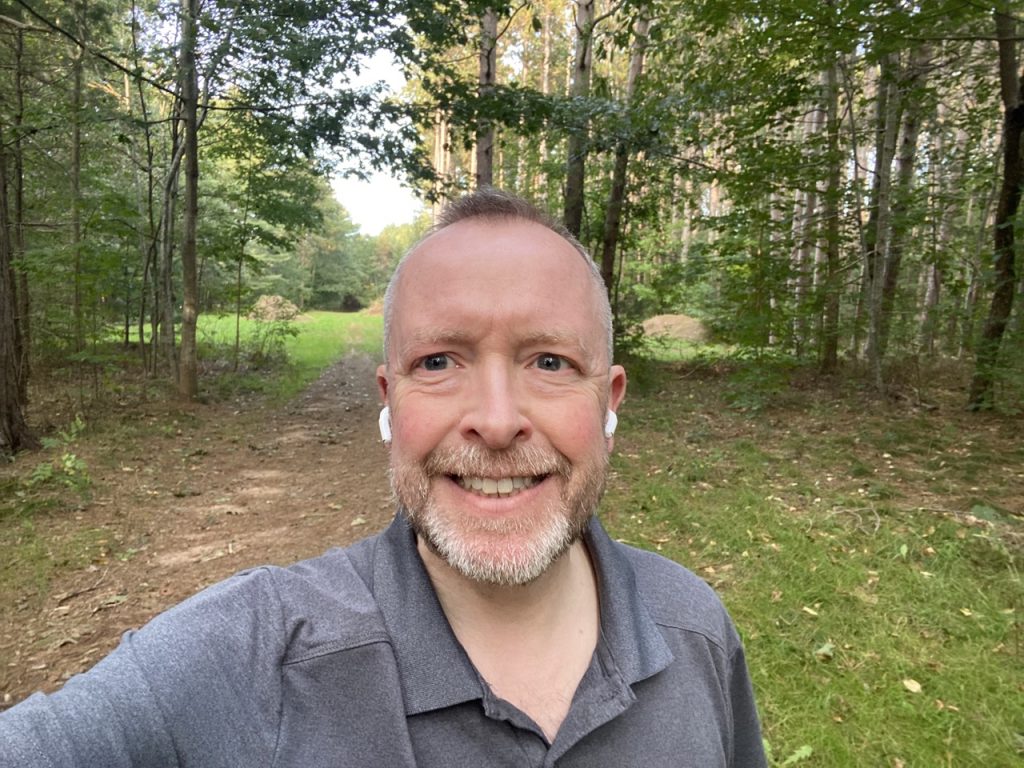 Matthew Guy, summer 2023, walking in the woods on a trail with EarPods. Smiling at the camera while taking a selfie. 