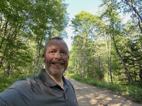 Middle aged white guy with short hair and AirPods on a trail with trees either side, looking roughly at the camera.