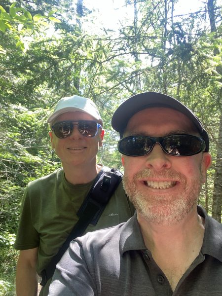 Two old white geezers posing for a selfie with sunglasses and baseball caps on, under the tree canopy.