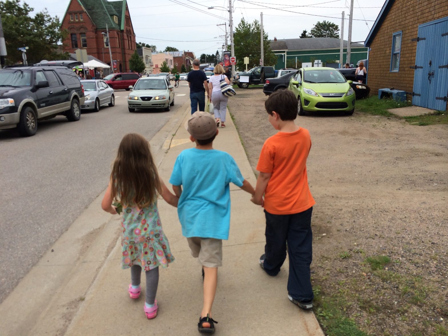 Three children from the back holding hands walking down a sidewalk