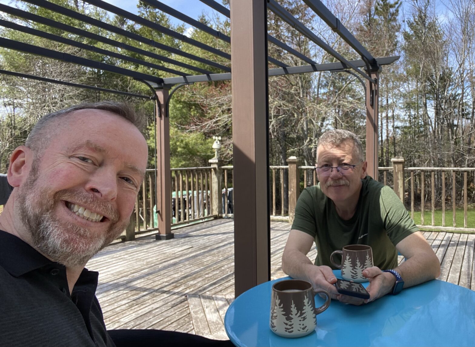 Two white guys in their 50s having coffee on a deck. Gazebo behind them. Sat at a blue metal patio table.